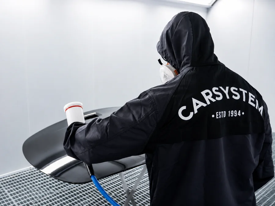 Lackierer im Carsystem-Overall trägt Klarlack auf ein schwarzes Bauteil in der Lackierkabine auf
